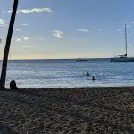 Afternoon beach scene with palm trees, boats, and swimmers in Hawaii at sunset.
